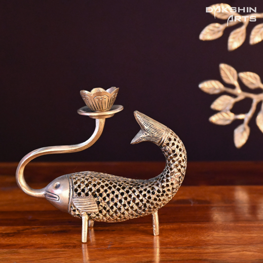 A brass candle holder shaped like a fish with intricate honeycomb detailing and a lotus-shaped top. The fish's mouth curls upward to support the lotus - Dakshin Arts