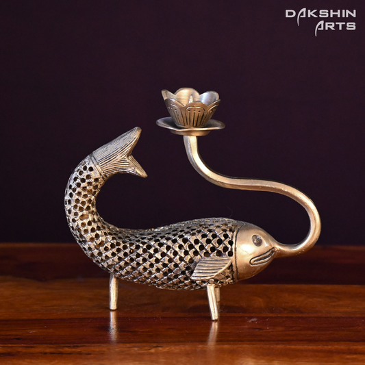 A brass candle holder shaped like a fish with intricate honeycomb detailing and a lotus-shaped top. The fish's mouth curls upward to support the lotus - Dakshin Arts