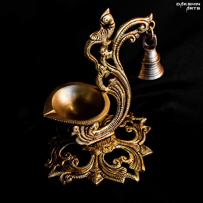 Ornate brass oil diya with intricate carvings, featuring a peacock design and a small hanging bell on a dark background - Dakshin Arts