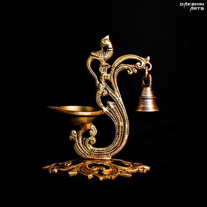 Ornate brass oil diya with intricate carvings, featuring a peacock design and a small hanging bell on a dark background - Dakshin Arts