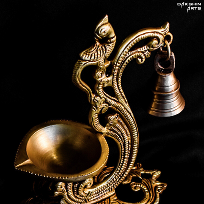 Ornate brass oil diya with intricate carvings, featuring a peacock design and a small hanging bell on a dark background - Dakshin Arts