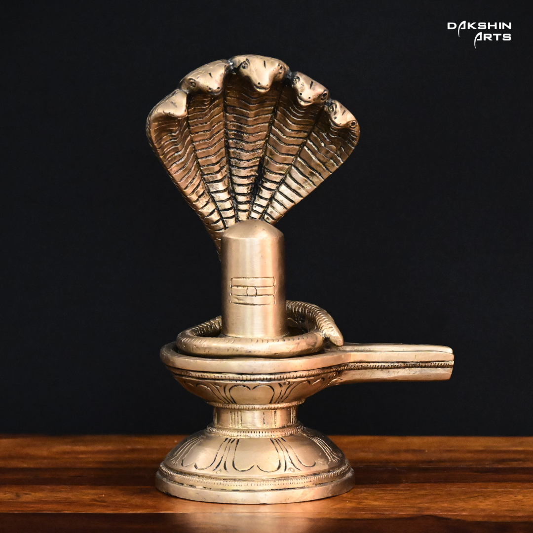 Brass sculpture of a Shivling with a five-headed cobra hood, set on a wooden surface against a black background- Dakshin Arts