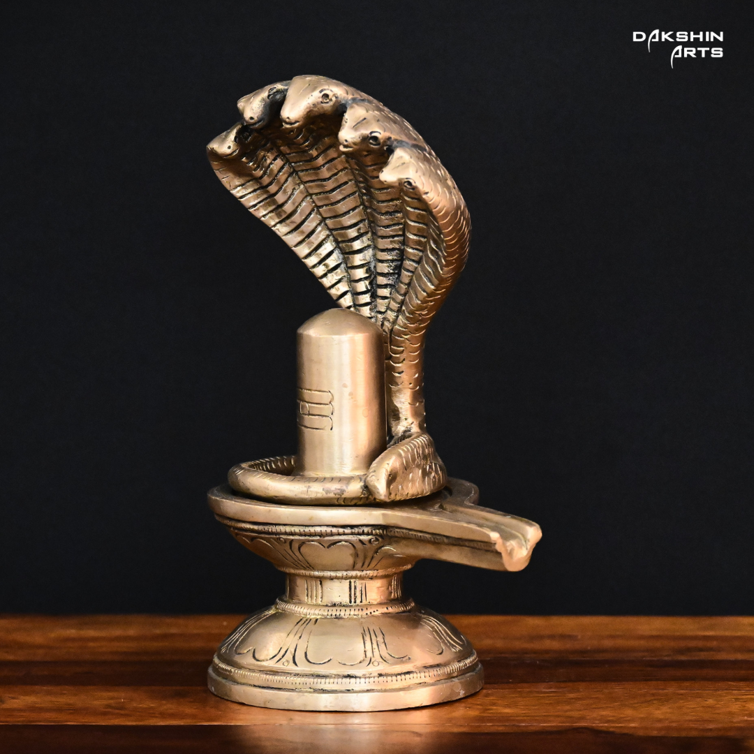 Brass sculpture of a Shivling with a five-headed cobra hood, set on a wooden surface against a black background- Dakshin Arts