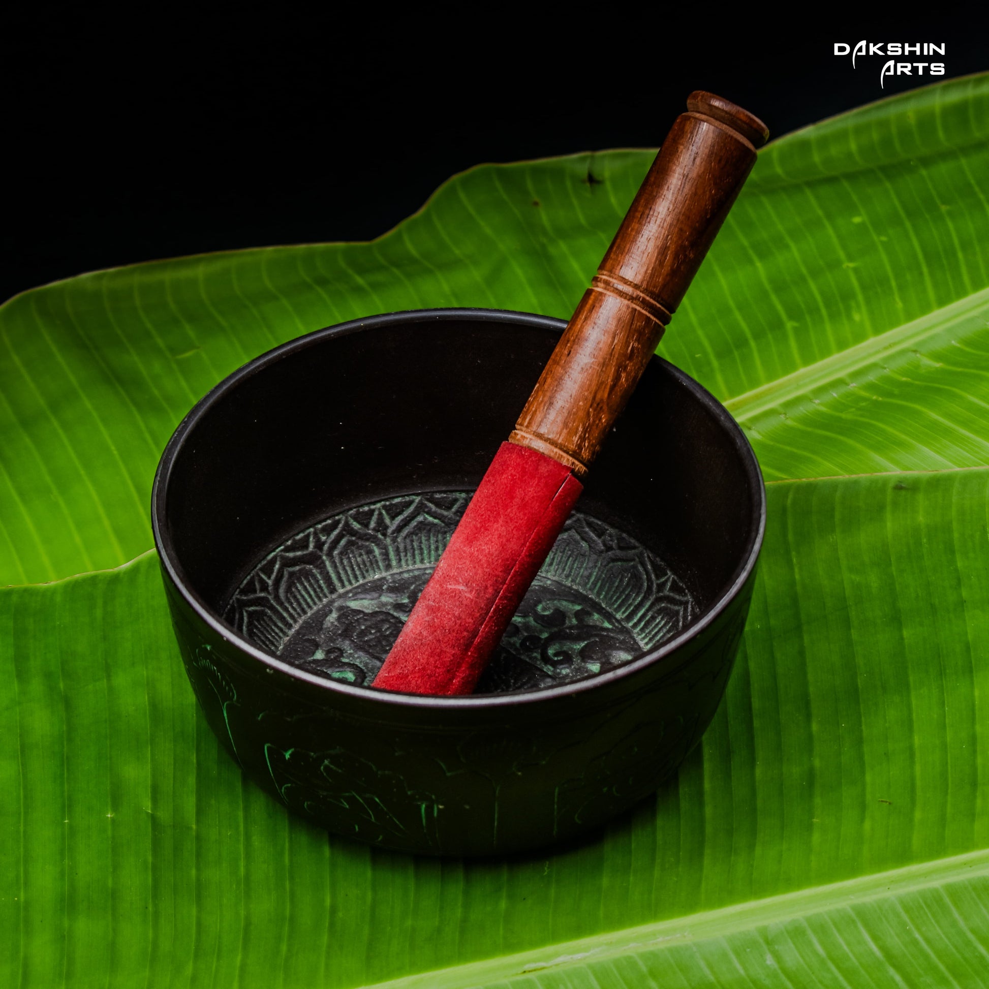 A dark singing bowl with intricate patterns sits on vivid green banana leaves. A red and brown mallet rests inside- Dakshin Arts