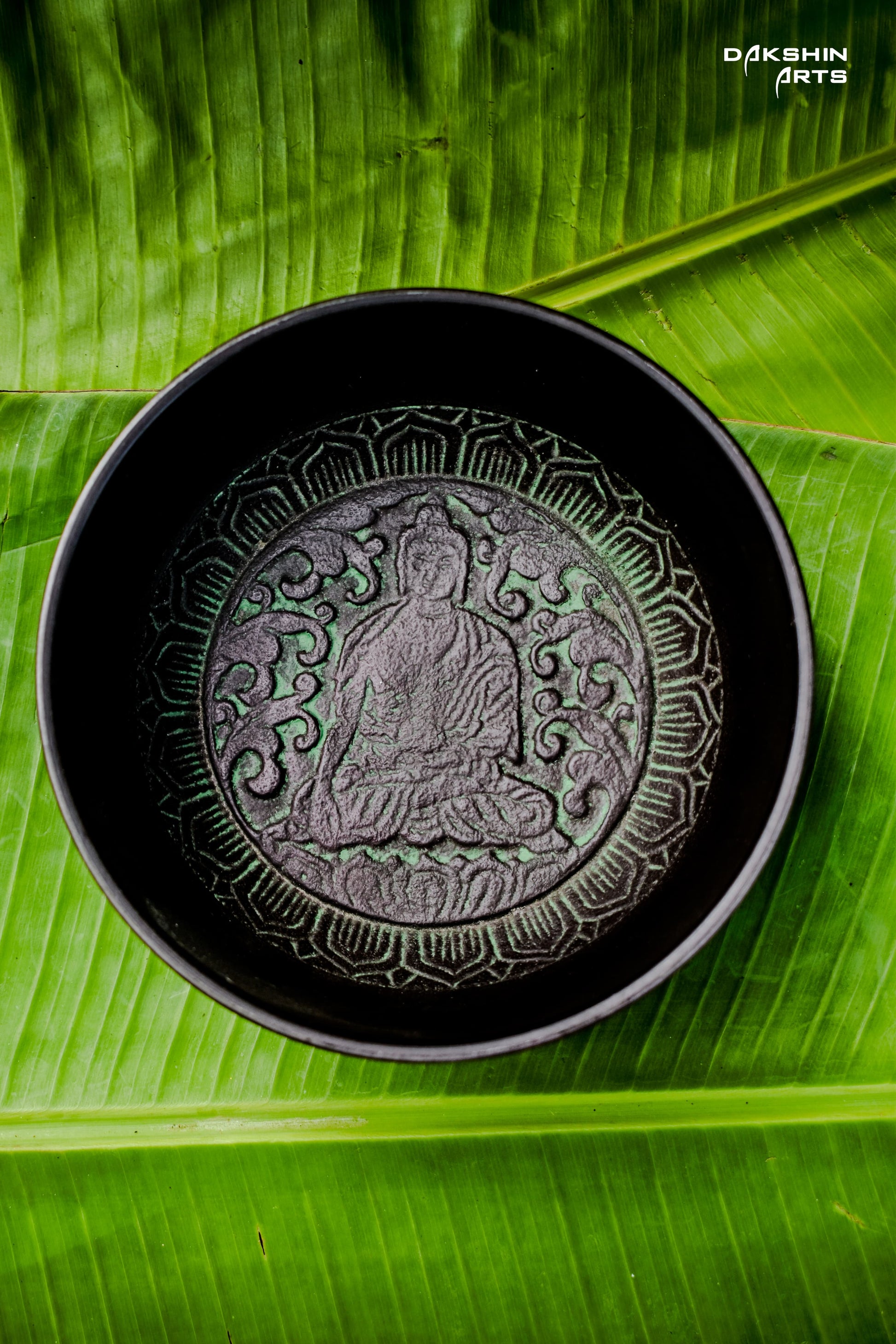 A dark singing bowl with intricate patterns sits on vivid green banana leaves - Dakshin Arts