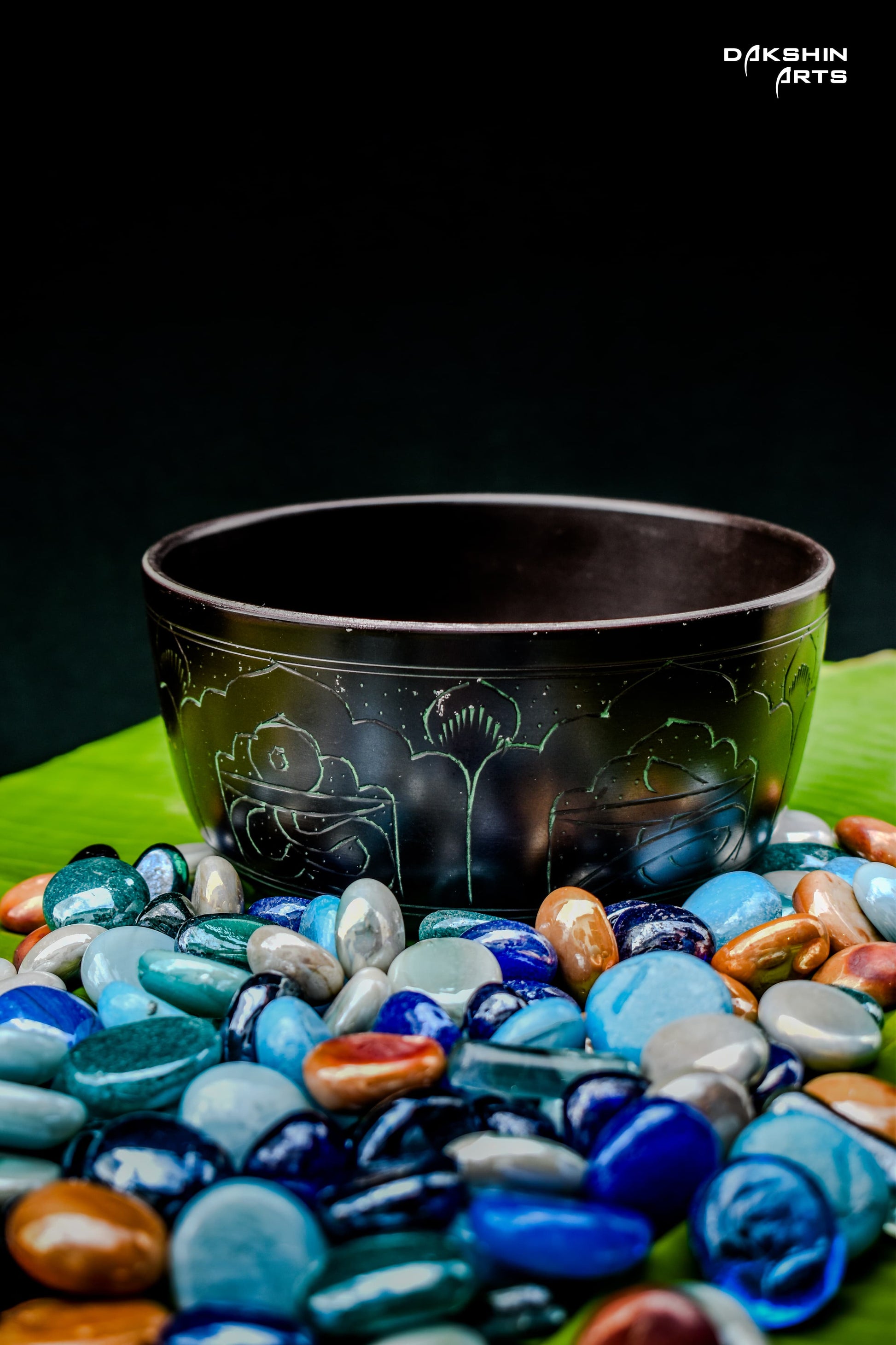A dark singing bowl with intricate patterns sits on vivid green banana leaves surrounded by pebbles - Dakshin Arts