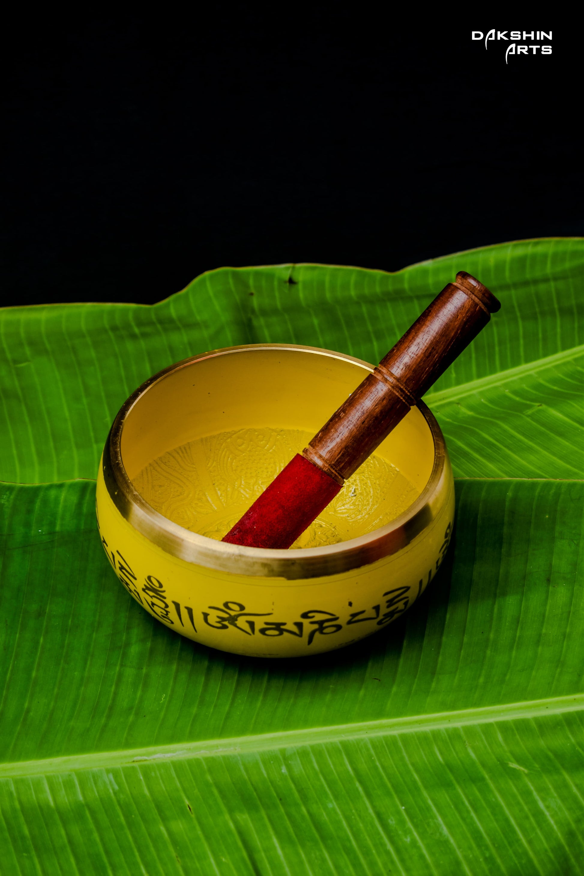 Yellow bowl with script sits atop  green  banana leaves, with a red mallet inside it - Dakshin Arts 