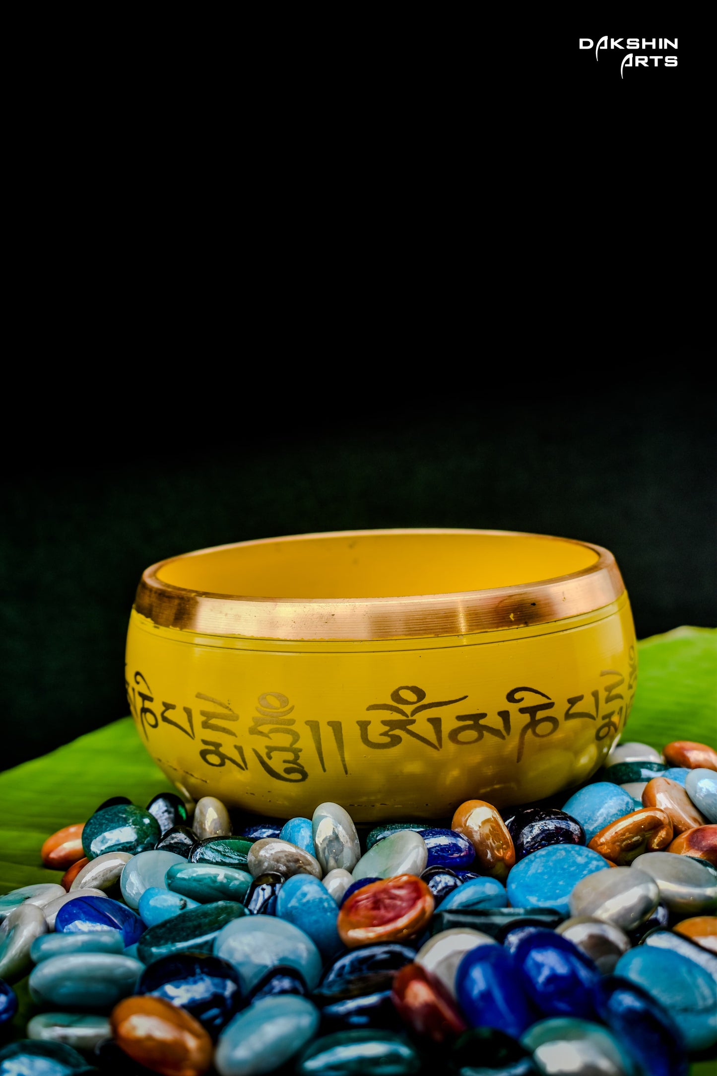 Yellow bowl with script sits atop colorful, polished stones on a green leaf- Dakshin Arts