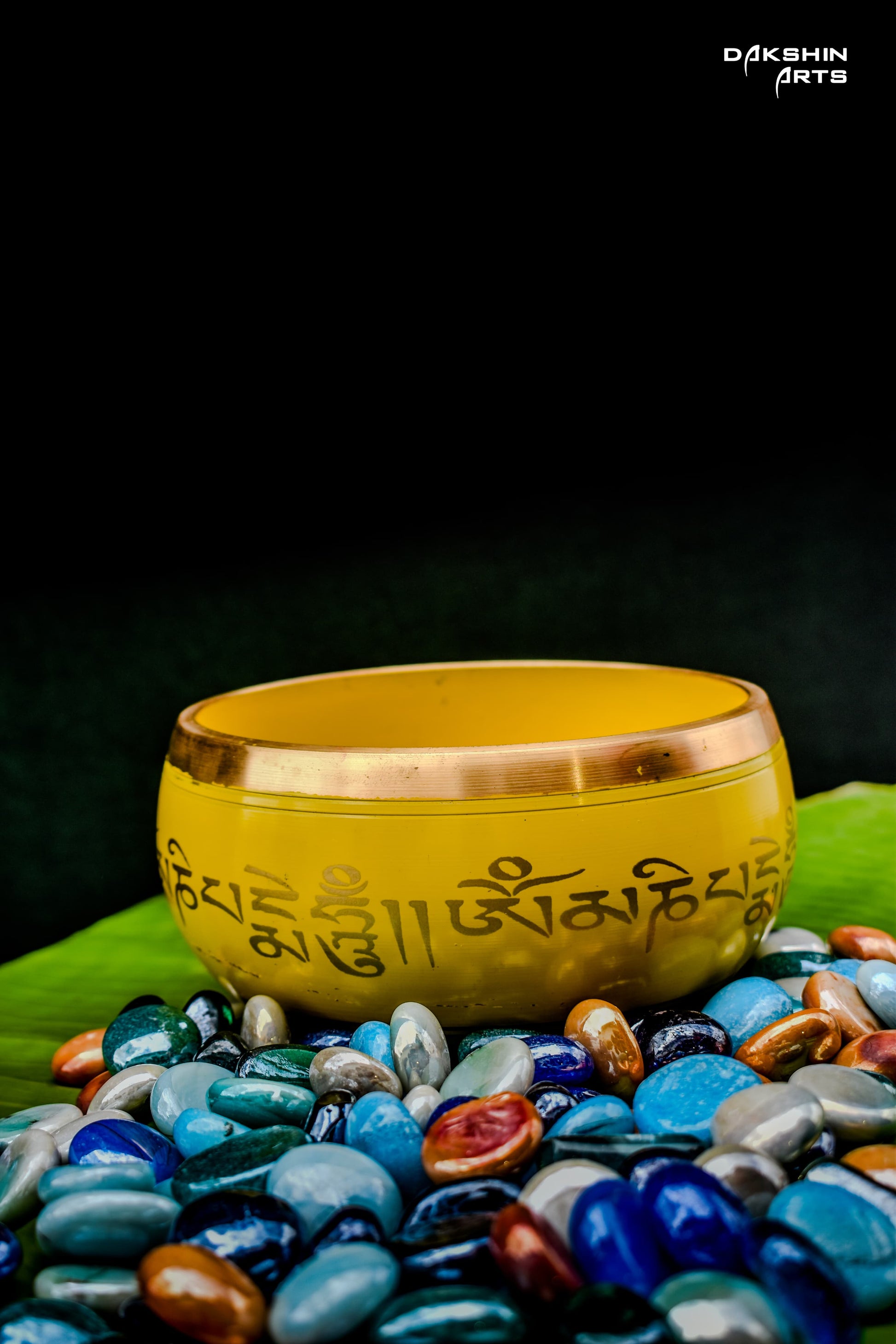 Yellow bowl with script sits atop colorful, polished stones on a green leaf- Dakshin Arts