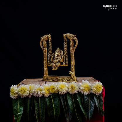 A brass Ganesha statue on a decorative swing with intricate bird carvings, adorned with flowers and green leaves- Dakshin Arts